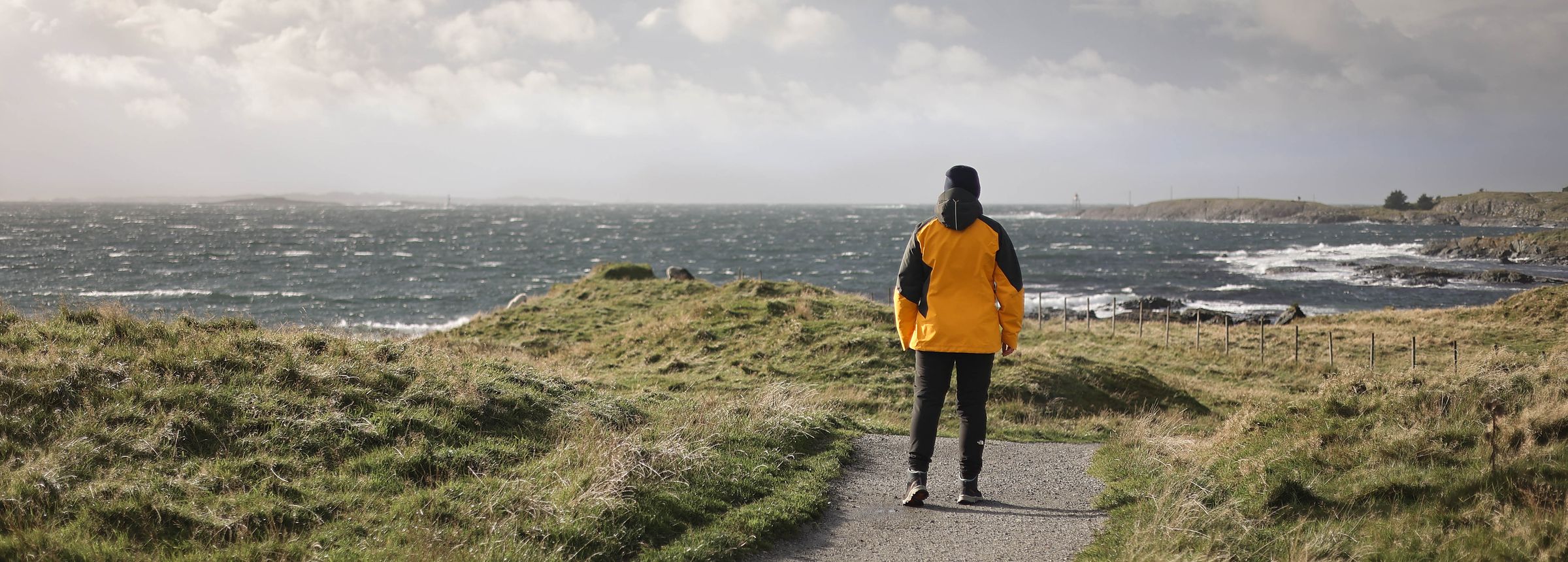 En person i gul jakke går bortover en grussti ut mot havet en vindfull høstdag. Graset ligger flatt, sjøen er hvit av vind, og lengst ute ligger lave skjær og en liten fyrlykt